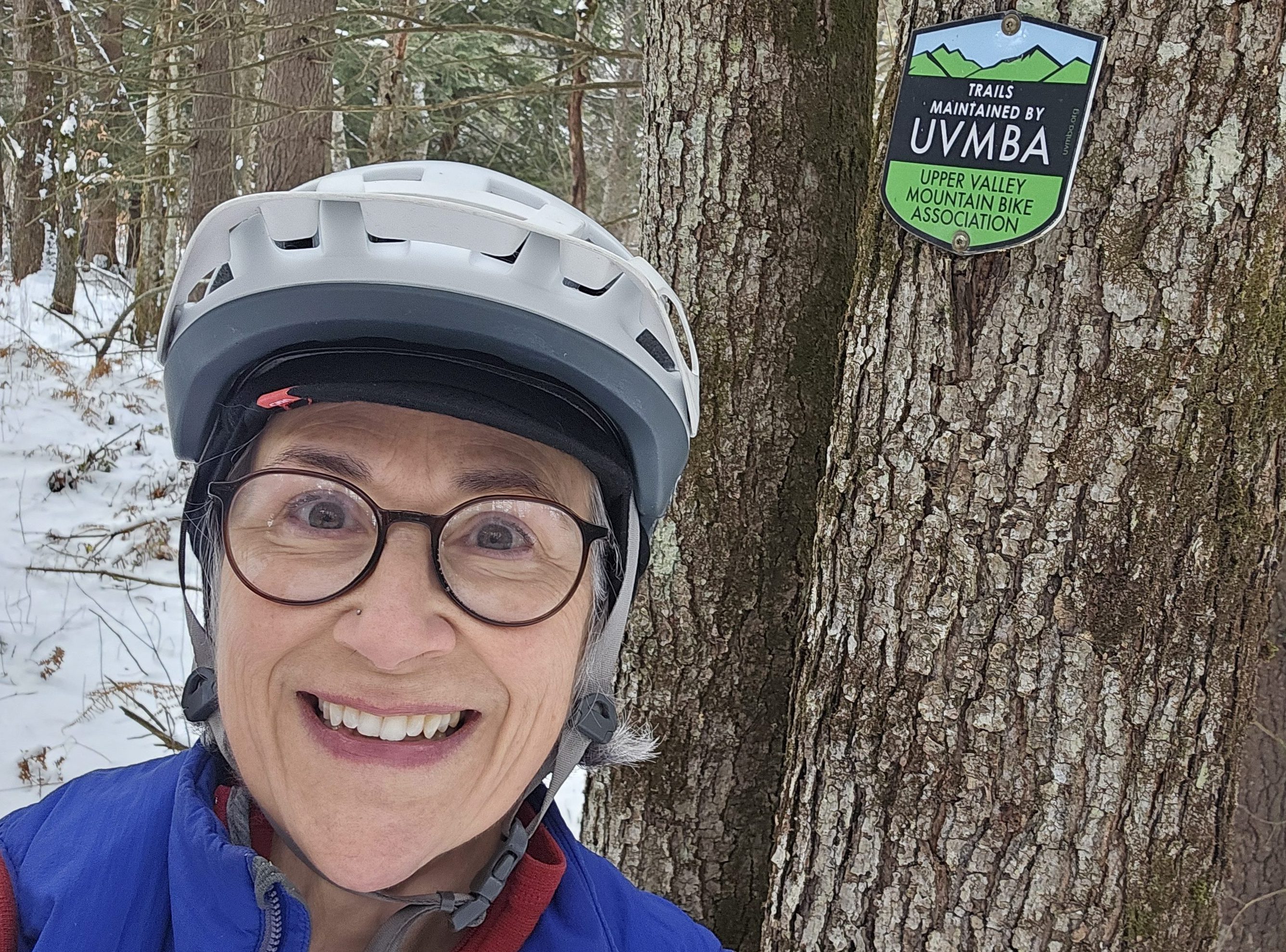 A smiling middle aged woman with a white bike helmet and round glasses, in front of a tree bearing a sign that says "trails maintained by Upper Valley Mountain Bike Association